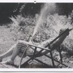 Black and white postcard depicting a photograph of Eudora Welty sitting outside by a sprinkler and a garden