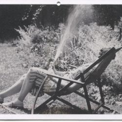 Black and white postcard depicting a photograph of Eudora Welty sitting outside by a sprinkler and a garden