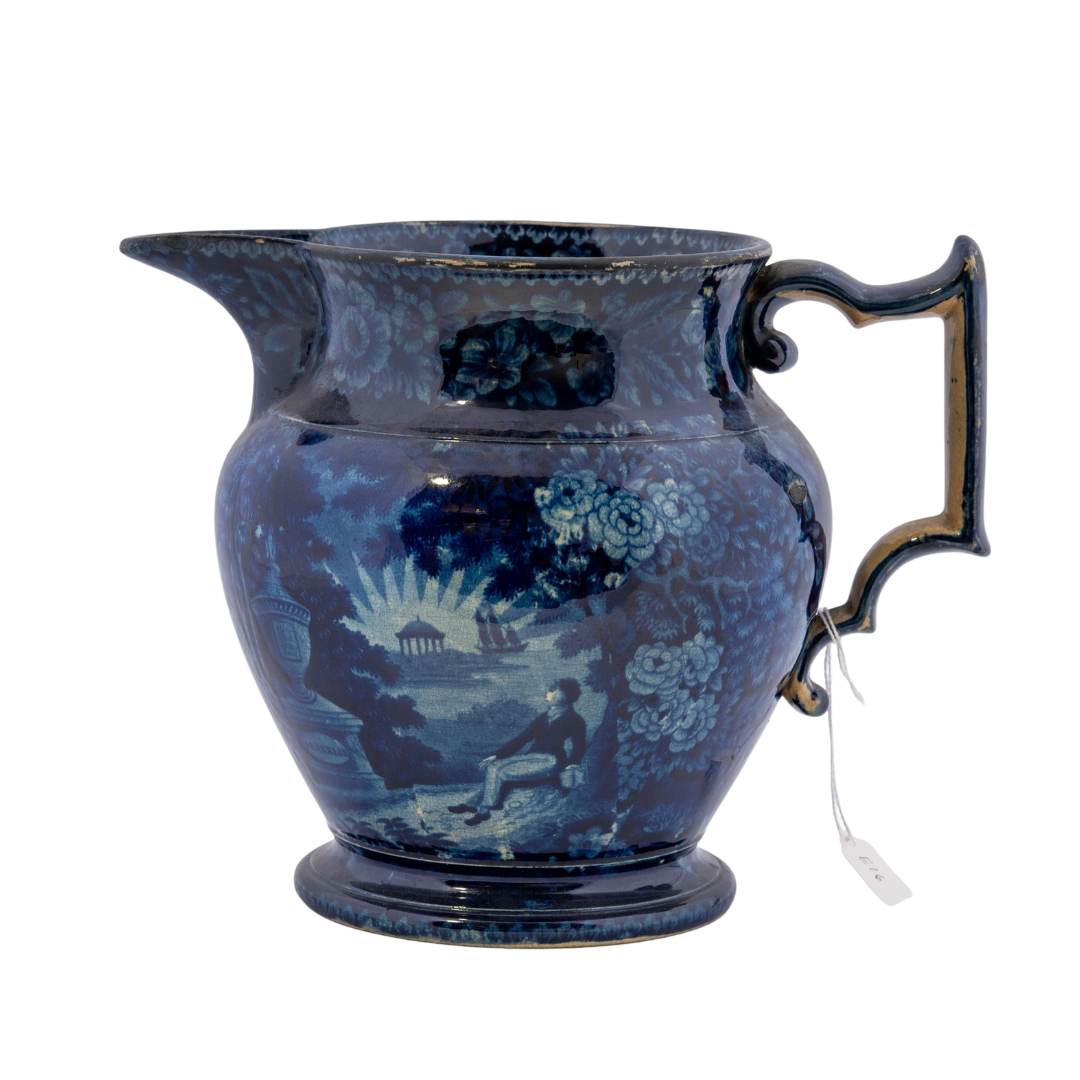 Blue Staffordshire ceramic transferware pitcher with blue glaze showing a scene of a man sitting across from a monument with the name "Franklin" at the base with a tall ship in the distance, known as "Lafayette at Franklin's tomb".