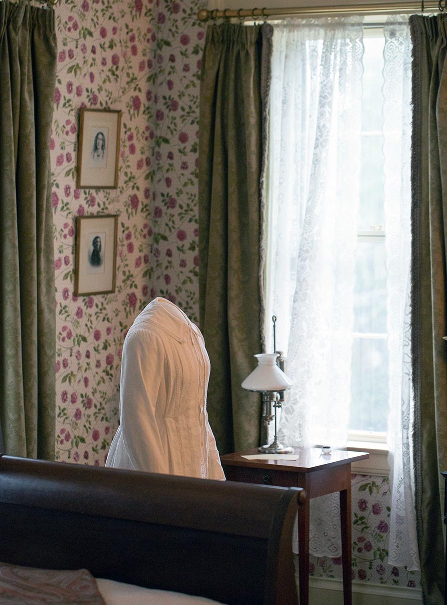 View of the bedroom with the bed in the foreground, and the desk and windows in the background. Between them stands a manikin with the poet's replica white dress