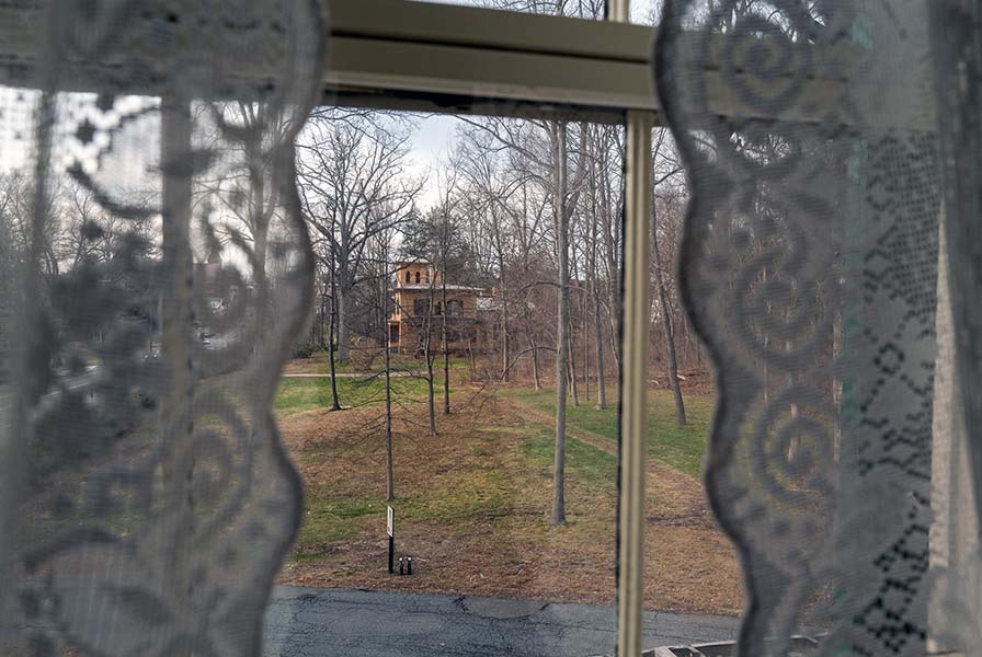 Paned window bordered by lace curtains. In view is a narrow path leading to a yellow Italianate house.