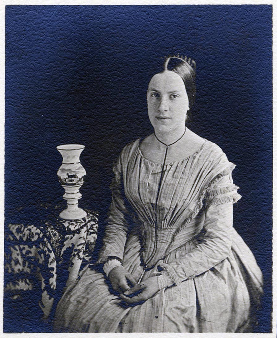 A young woman in a dress with a fan-pleated bodice sits with her hands folded in her lap. 