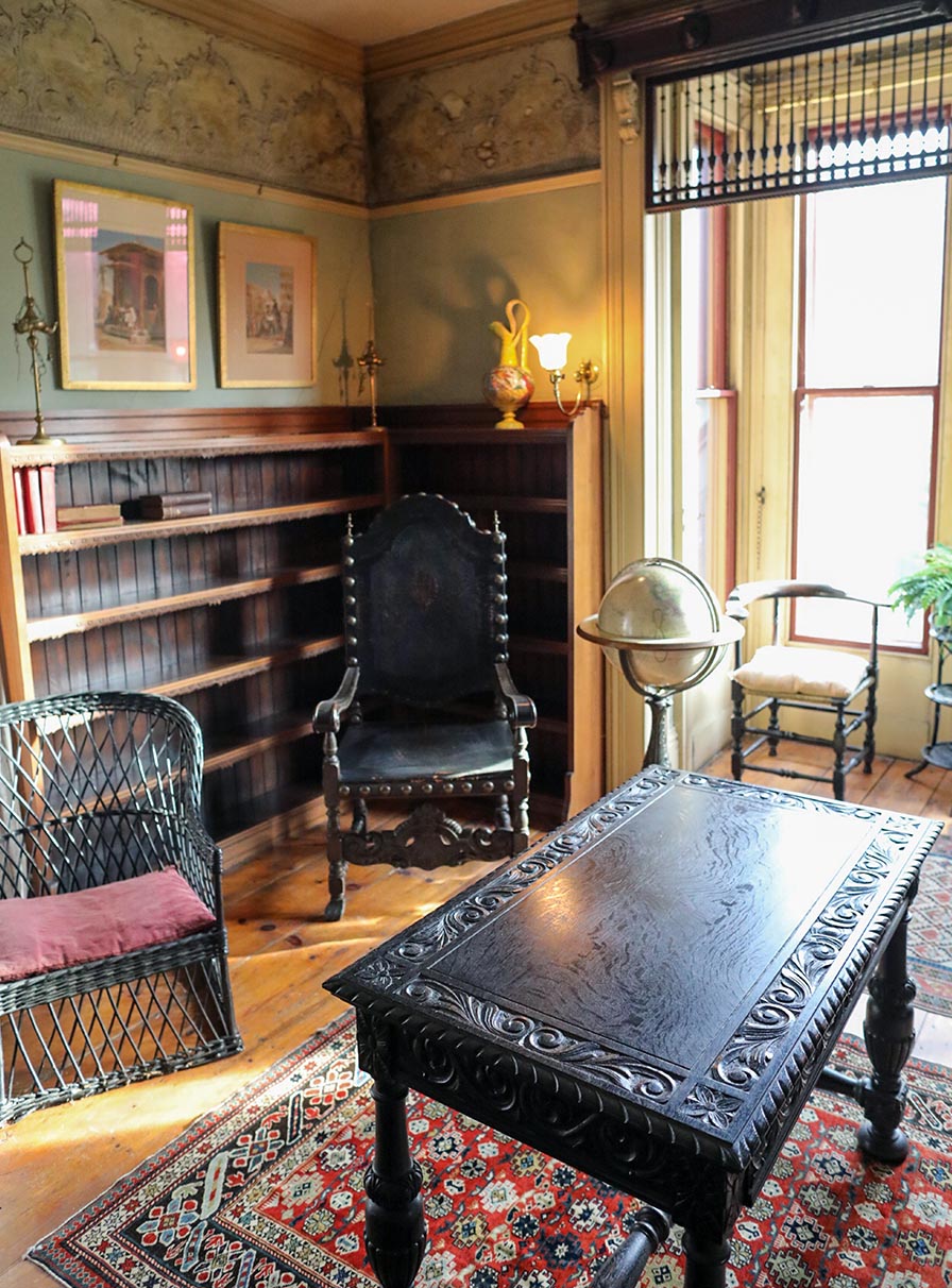 Library with green papered walls with an elaborate frieze. In center is a renaissance revival table with carved top and sides