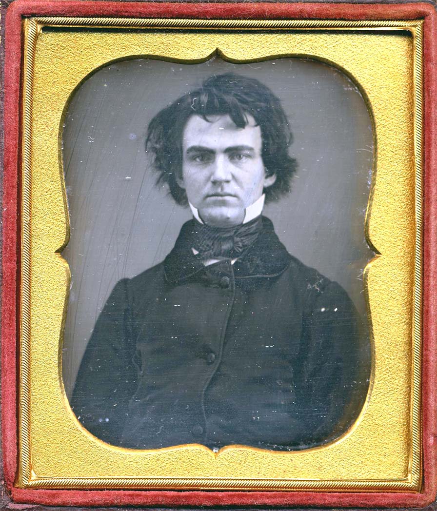 A young man wearing a jacket, cravat, and high starched collar. His hair is strikingly messy