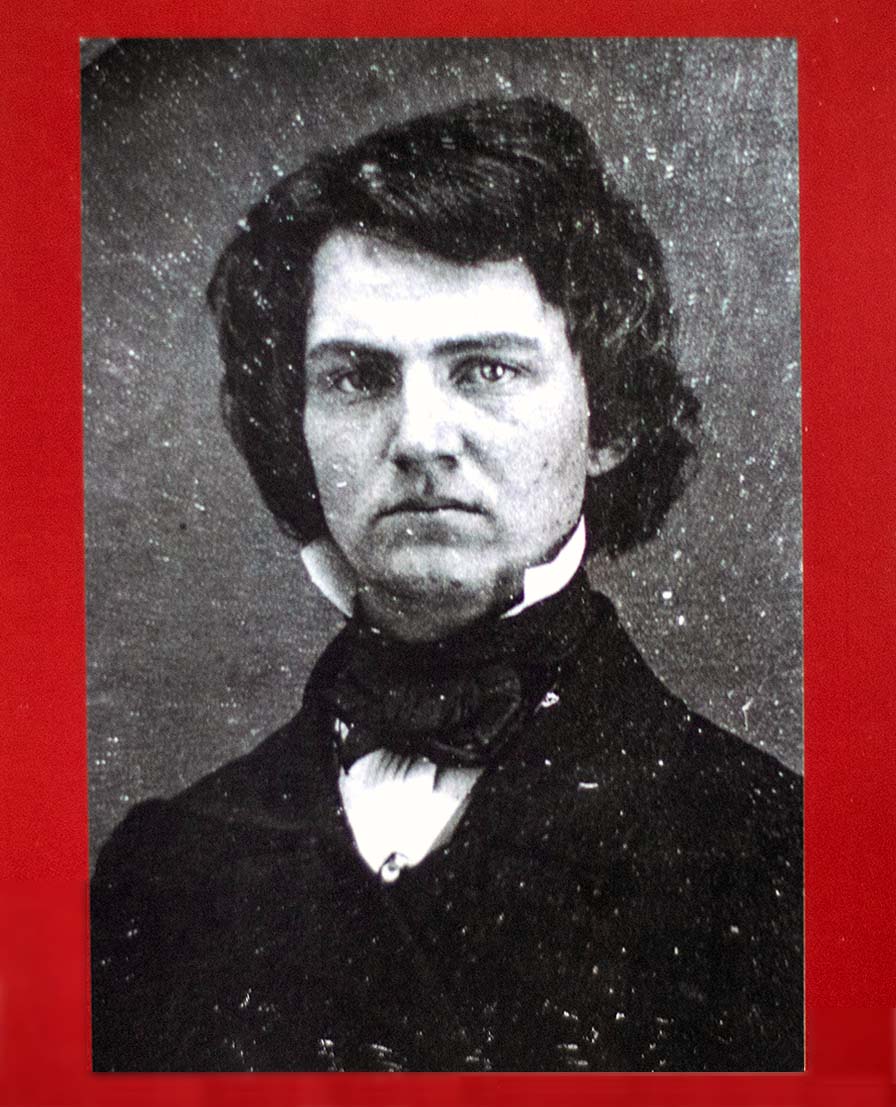 A serious young man with thick hair wears a dark coat and upright white collar with necktie.