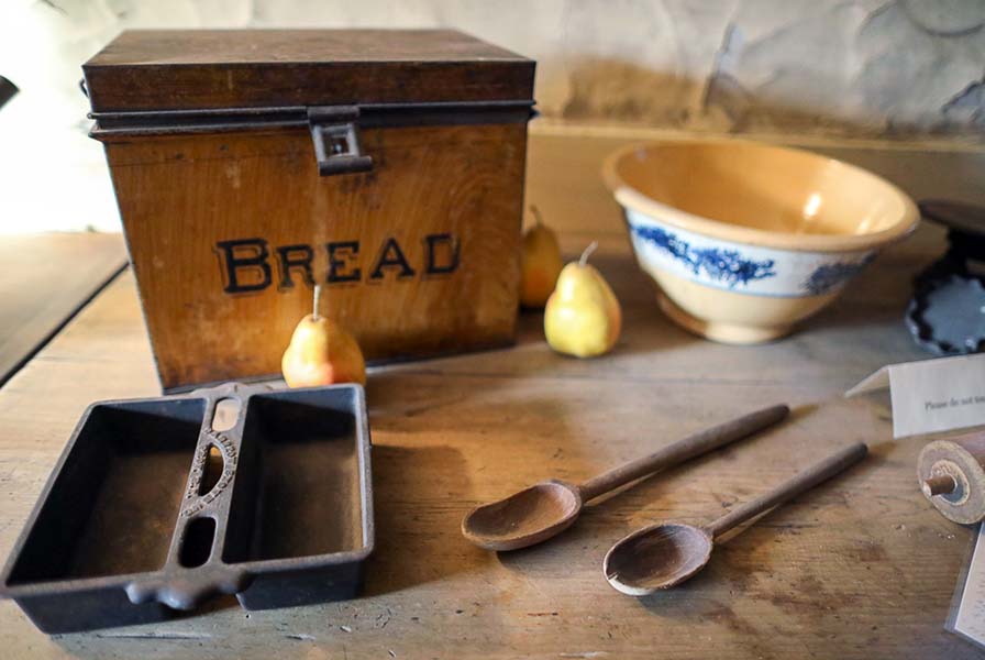 Tin bread box surrounded by pears, twin loaf pan, yellow ceramic bowl, rolling pin, and two spoons rest on weathered table.