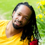 picture of Jericho Brown: a black man wearing a yellow t-shirt smiles in front of some daffodils