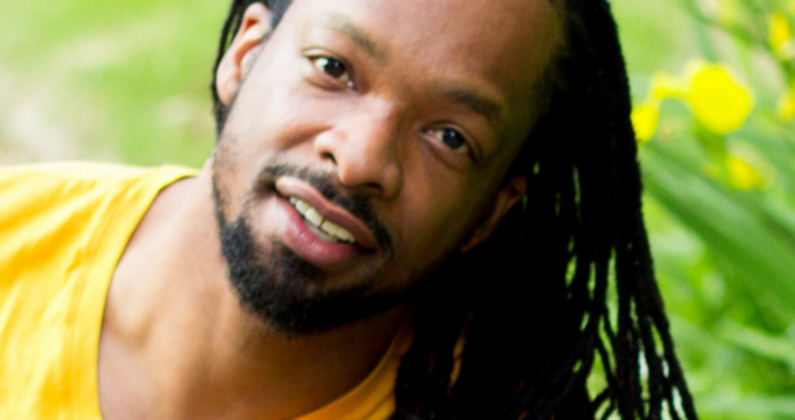 picture of Jericho Brown: a black man wearing a yellow t-shirt smiles in front of some daffodils