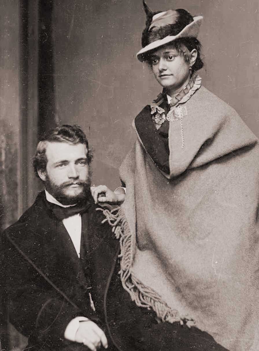 Black-and-white photo of a young woman in a feathered hat resting her hand on the shoulder of a seated man in a suit. 