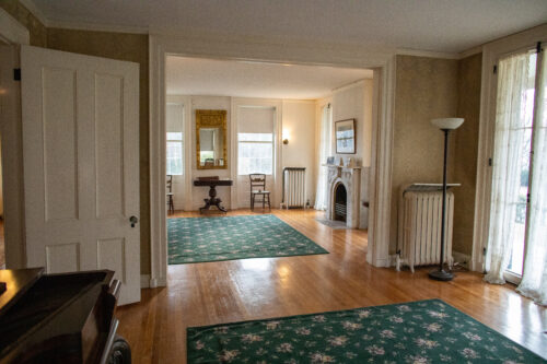 The parlor at the Homestead. Wide-shot featuring a doorway, two chairs, a desk, a gold framed mirror, and the fireplace