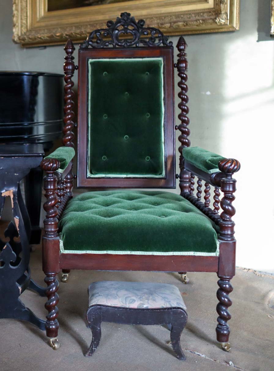 A wooden armchair with green velvet upholstery and an elaborately carved crest. The arms, back, and legs are spiral-turned.