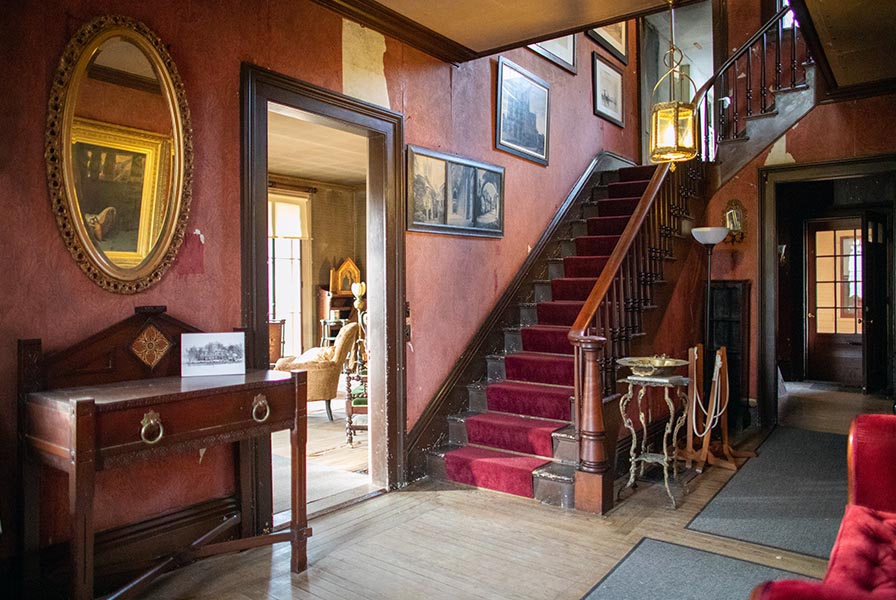 Entry hall with staircase, finished in rich reds and woods. There is a calling card receiver & round gilt mirror.