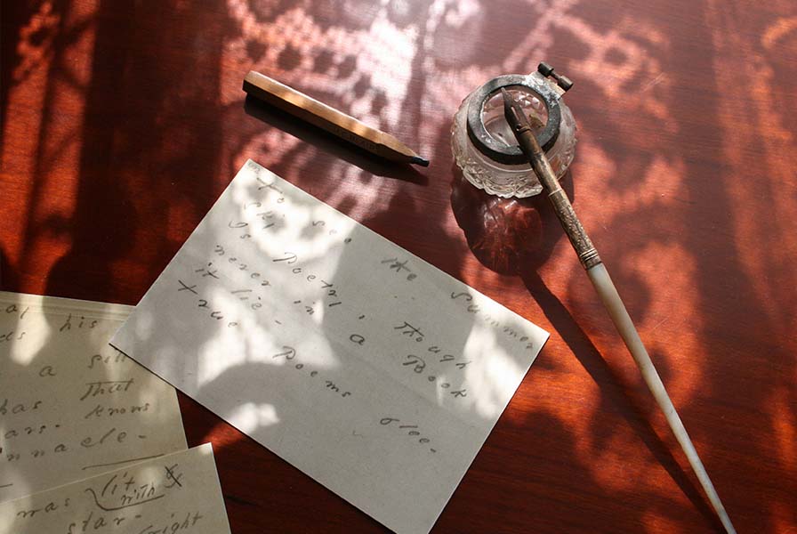 Wood surface cast in lacy shadows. On top: a dip pen resting on an inkwell, a pencil stub, and verse printed on paper scraps.