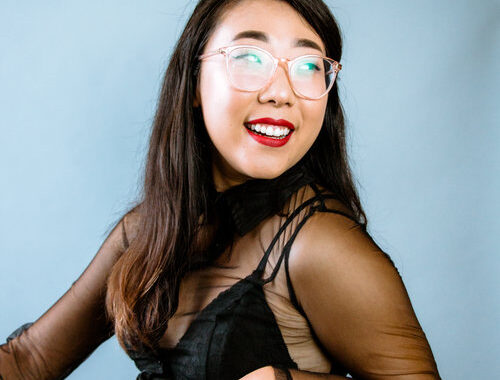 picture of Franny Choi, dressed in black, leaning back in a chair. Franny has long black hair, clear glasses, and bright red lipstick and is smiling
