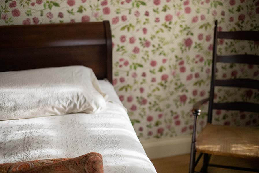 A sleigh bed with white coverlet next to a ladder-backed rocking chair. Scattered sunlight warms the scene.