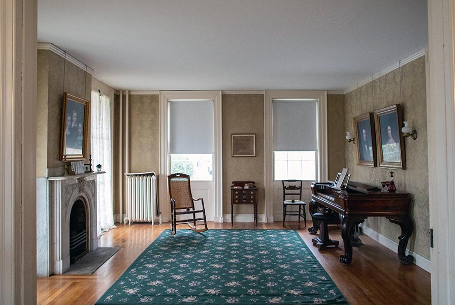 Two windows look north; at right an antique piano with two portraits above, at left a decorative marble fireplace with chairs beside.
