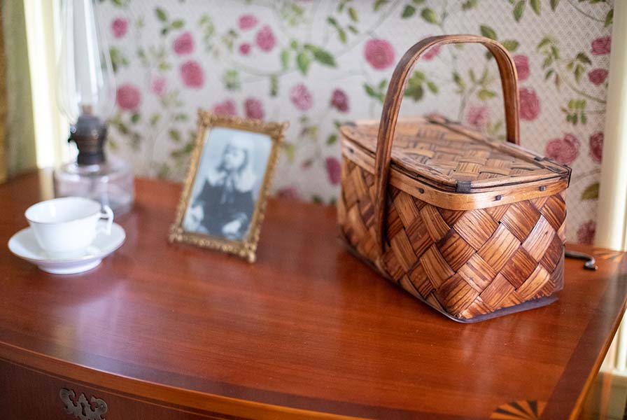 Woven lidded basket with handle resting on a wooden bureau next to a teacup, oil lamp, and framed photograph of a child.