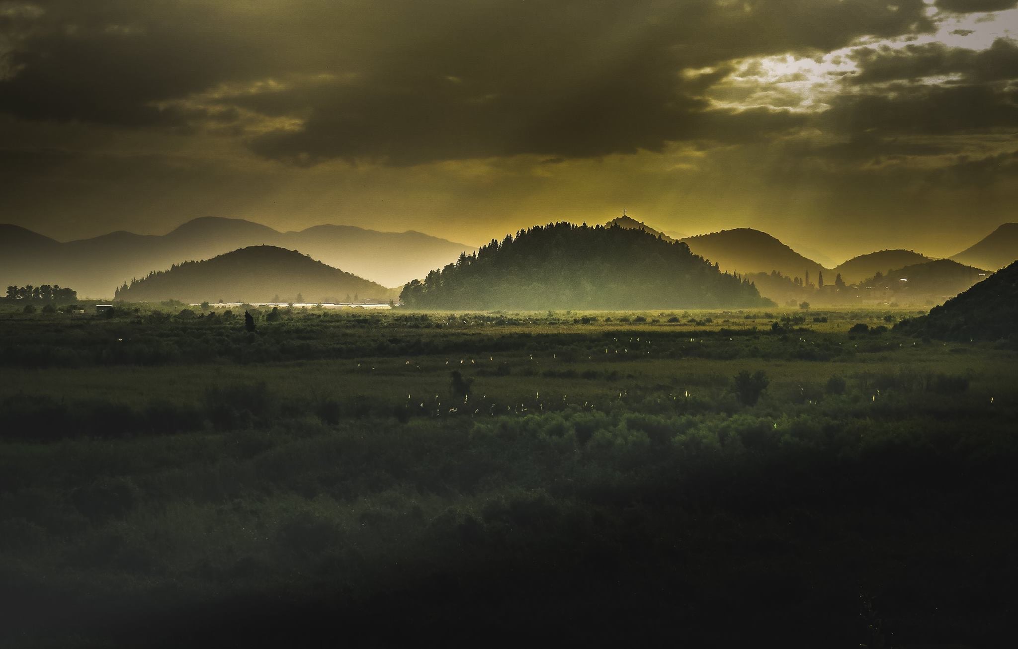 A photo taken from the perspective of a cemetery in Bosnian, featuring rolling, misty hills on a thickly cloudy day; In the foreground is a seemingly endless expanse of field. The whole photo is in tones of deep yellows, greens and blacks.