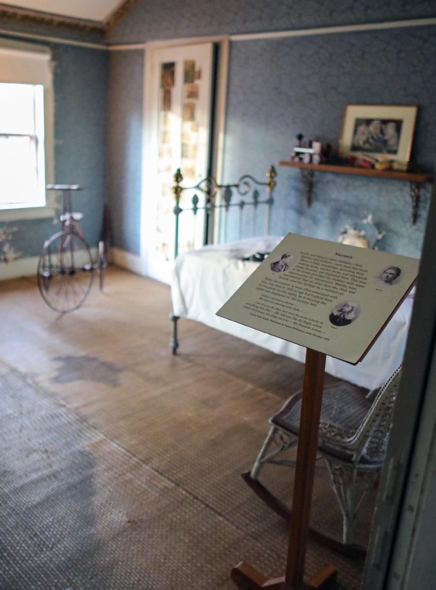 Doorway view of a room with blue wallpaper and straw floor matting. A sunlit window illuminates a twin bed and velocipede.