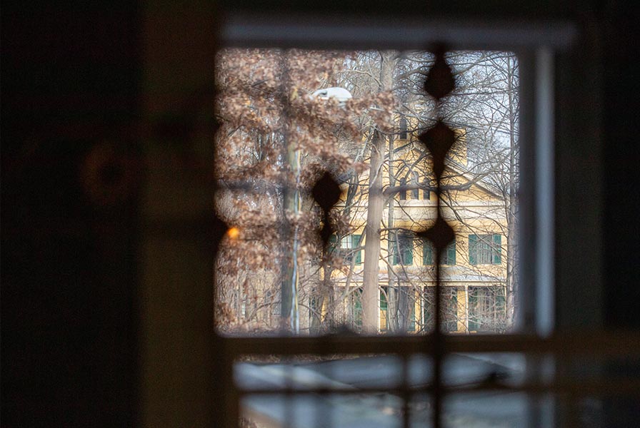 An autumn scene of the Homestead, seen through a window. There are shadows of a metal bed frame in the foreground.
