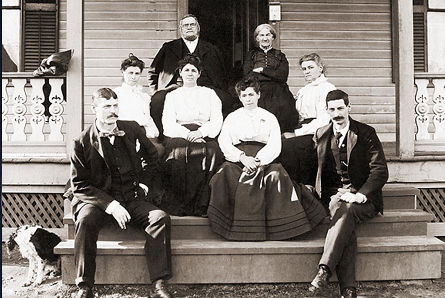 5 women and 3 men in formal attire seated on the porch and front steps of a wooden house. In the bottom left is a small dog.