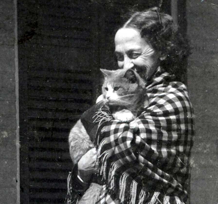 A smiling woman stands, hugging a tabby cat to her chest. She wears a checkered shawl and full skirt.