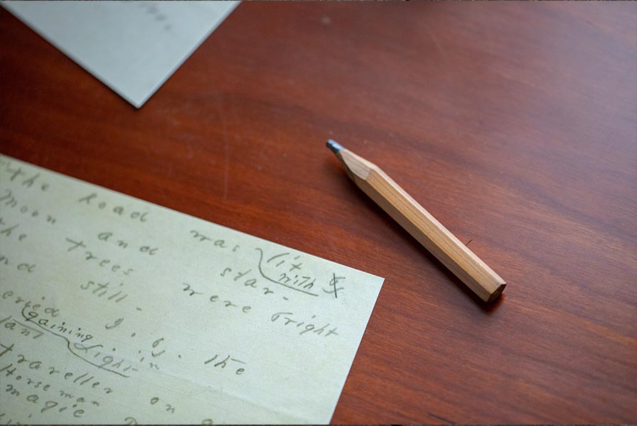 A corner of a Dickinson manuscript on the desk shows her marks indicating variant word choices; a short stubby pencil lays beside.