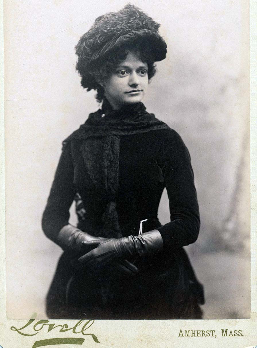 A fashionable young woman with curly hair, velvety dress, and feathered bonnet stands with gloved hands lightly folded.