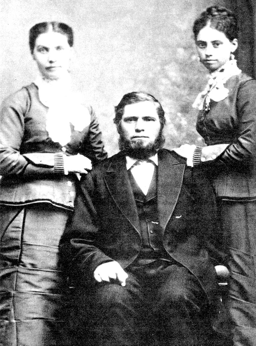 Maher, a woman in a jacket and skirt, stands to the right of a seated man in a suit, and a young woman with braided bun.