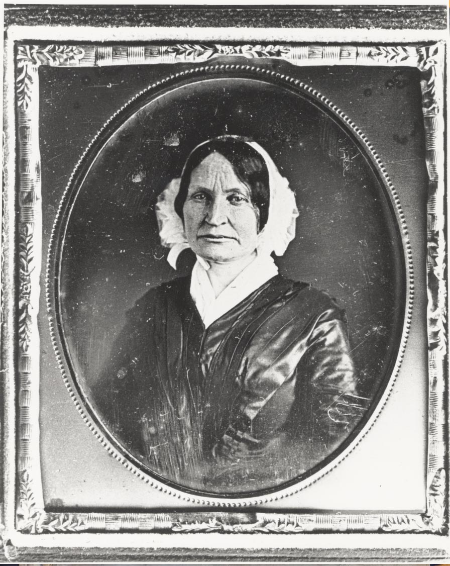 Worn daguerreotype of mature woman with creased forehead in a dark, pleated dress with white ruffled bonnet.
