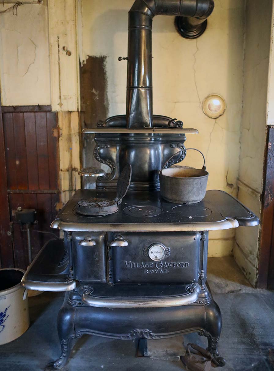 A large black cast iron stove stands against an old plaster wall. On top rest heavy iron pots and pans.