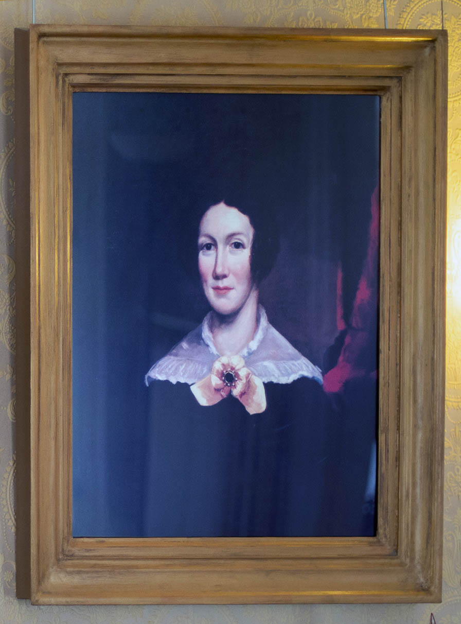 Oil portrait of a young woman. Slight smile, lace around her shoulders pinned with a yellow ribbon.