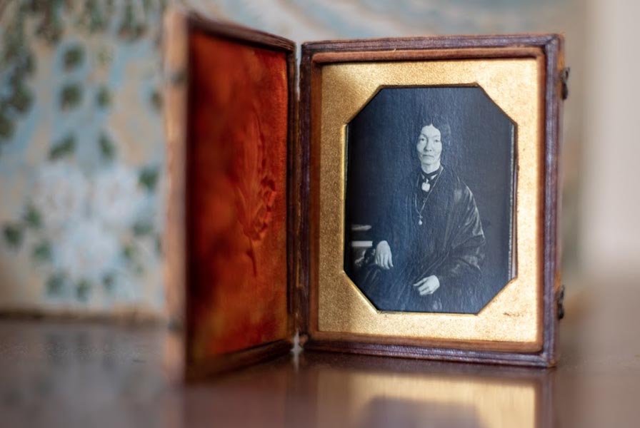 Daguerreotype in case of a seated woman in a dark dress and shawl. The case rests open on a table in front of blue bandbox.