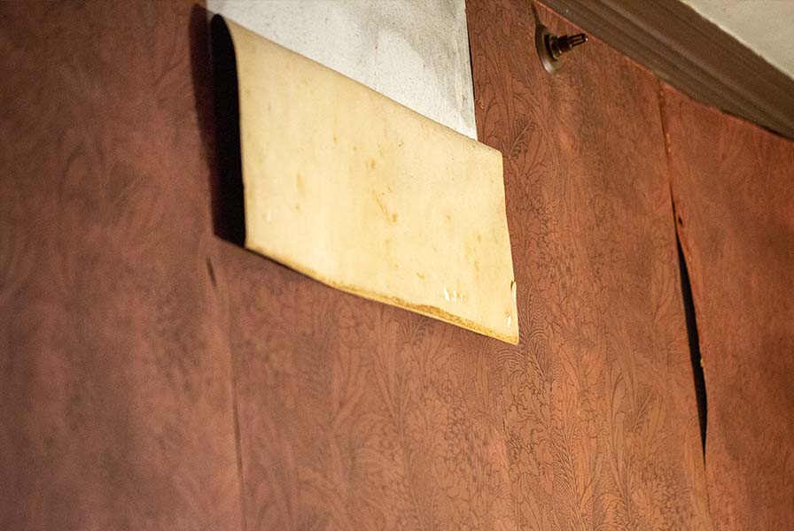 Faded burgundy wallpaper with ornate floral figures, loosening with age. One strip hangs from the wall, exposing the plaster