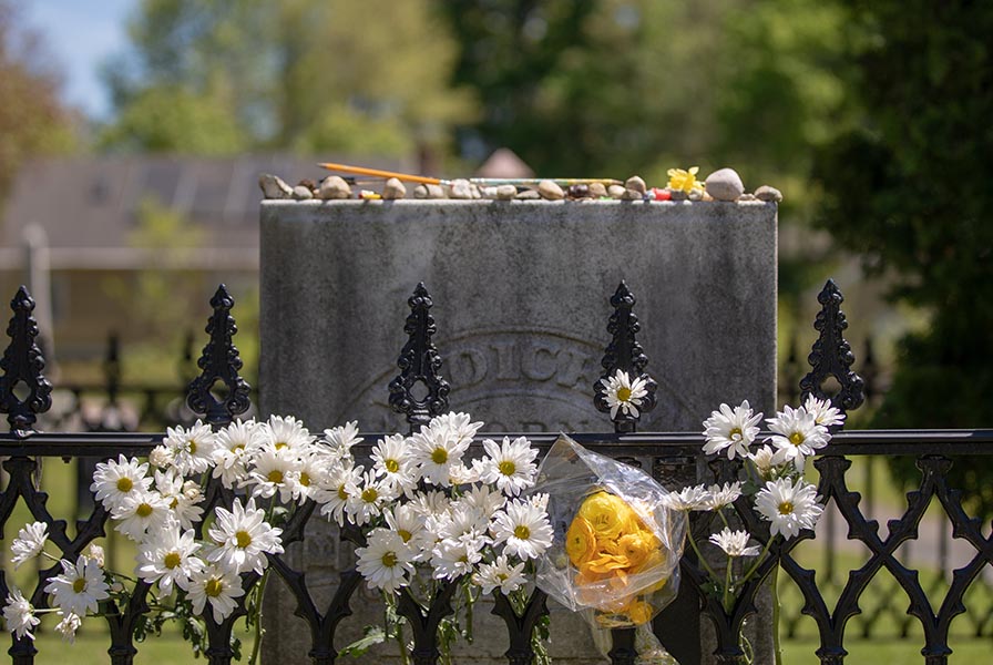 A granite headstone behind a wrought iron fence bedecked with flowers. Pencils and other offerings sit atop.