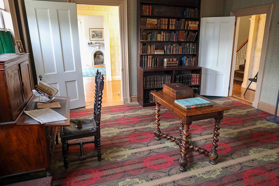 Carpeted library finished green, red, and tan with tall, full bookshelf, secretary desk, and library table with turned legs.