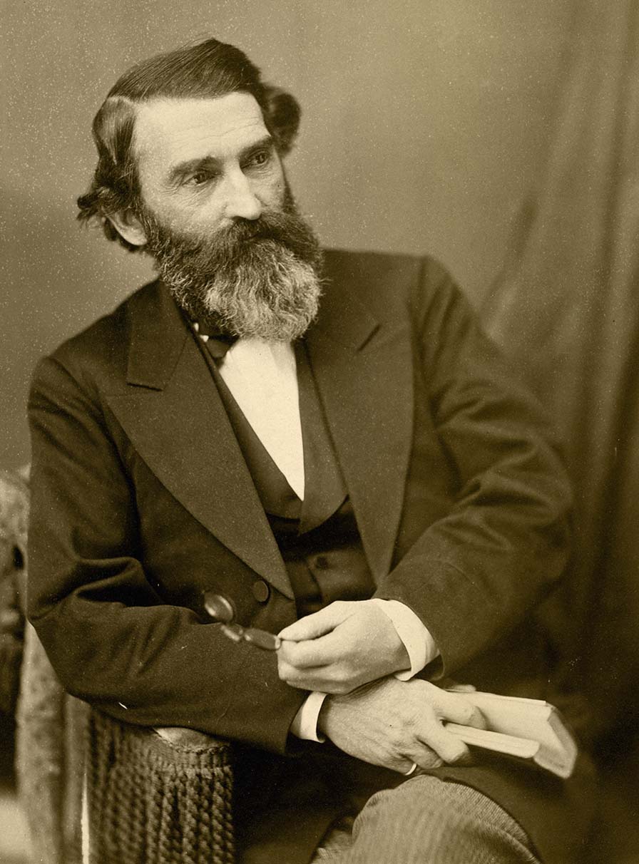 A seated man with a full beard and mustache. His arms are crossed. He holds a book in one hand and glasses in the other.