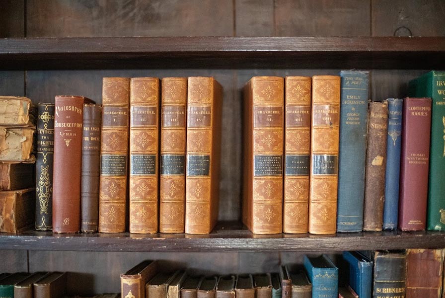 Bookshelf with a handsomely gilded 8 volume set of Shakespeare in center. Vol. 5 has been removed from the shelf.