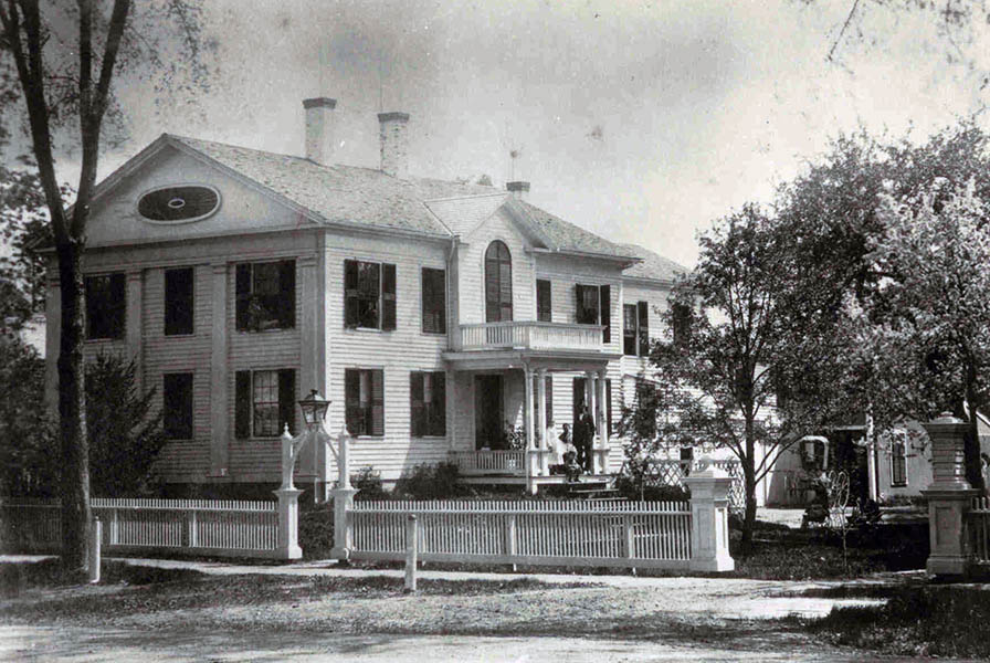 Black and white photo of a large white house with dark shutters. A striking fence lines the sidewalk.