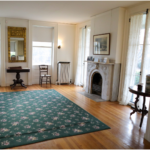 The homestead parlor with 4 windows, a fireplace, two chairs, a rug and a side table.