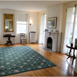 The homestead parlor with 4 windows, a fireplace, two chairs, a rug and a side table.