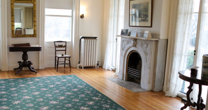 The homestead parlor with 4 windows, a fireplace, two chairs, a rug and a side table.