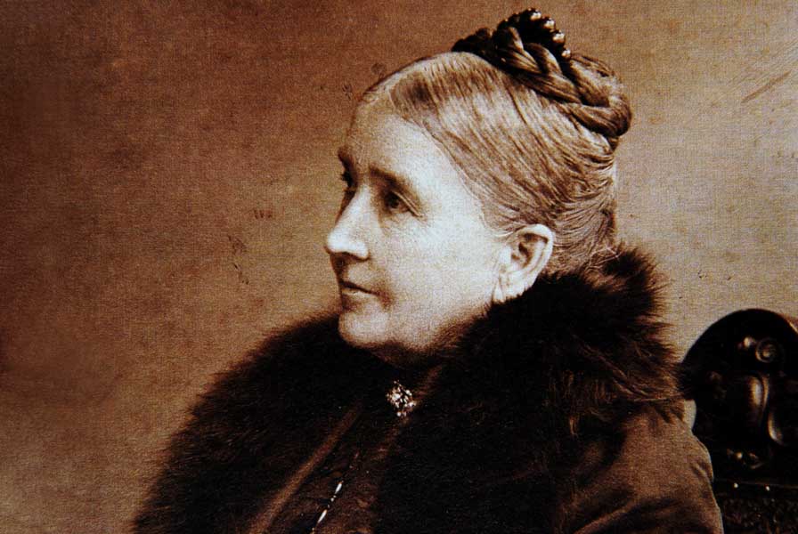 A mature woman with a high, braided updo sits in profile. She wears a fur-trimmed coat.