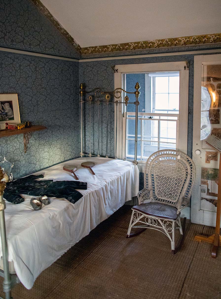 Nursery room corner with a narrow brass bed and wicker rocking chair. Behind the bed, an interior window faces the hallway.