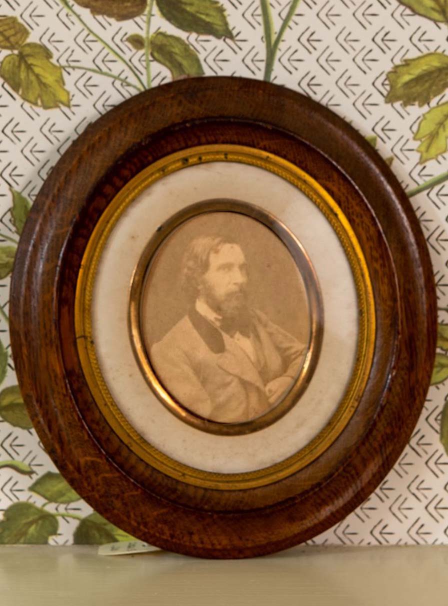 Framed faded photograph of a bearded man in a light-colored suit. The oval wooden frame has a gold metal inner border.