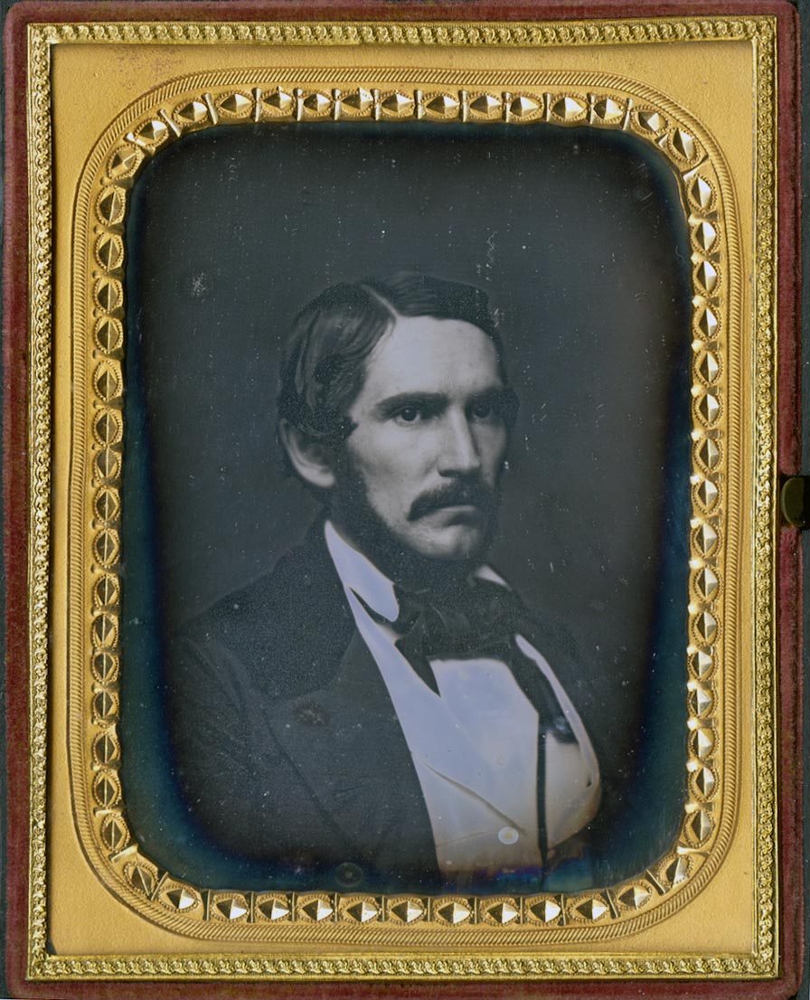 Daguerreotype of a man with mustache, long sideburns, and strong features wearing a suit with white waistcoat. In gilt case.