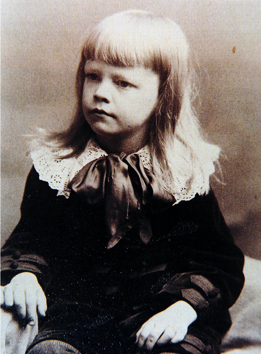 Historic photograph of a young boy with long, fair hair and bangs. He wears a velvet suit with a large lace collar and bow.