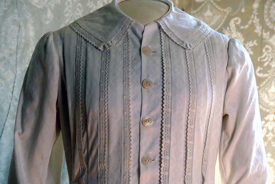 Front of a collared white cotton pique wrapper trimmed with rows of lace and mother-of-pearl buttons.