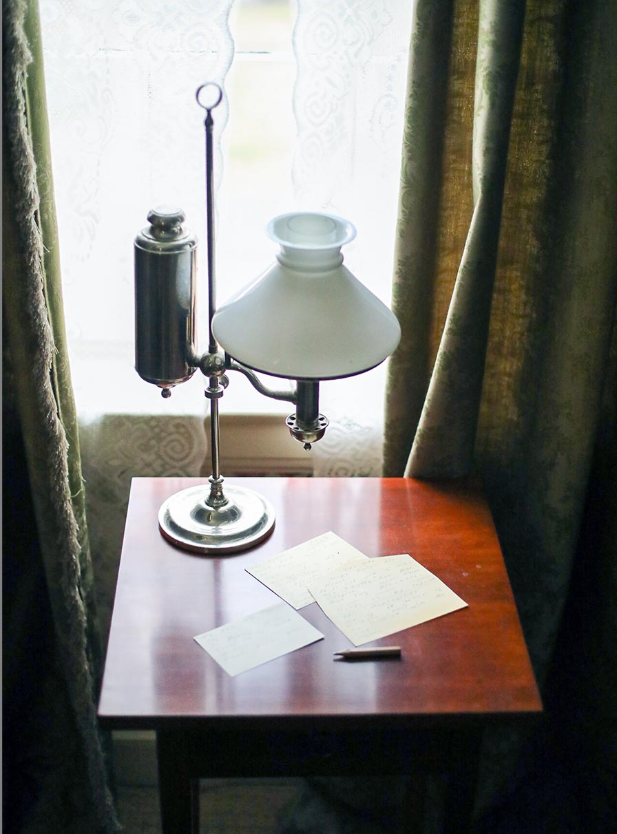 Small cherry table with square top in front of a window. On table: student lamp with large oil tank, pencil nub, paper scraps.