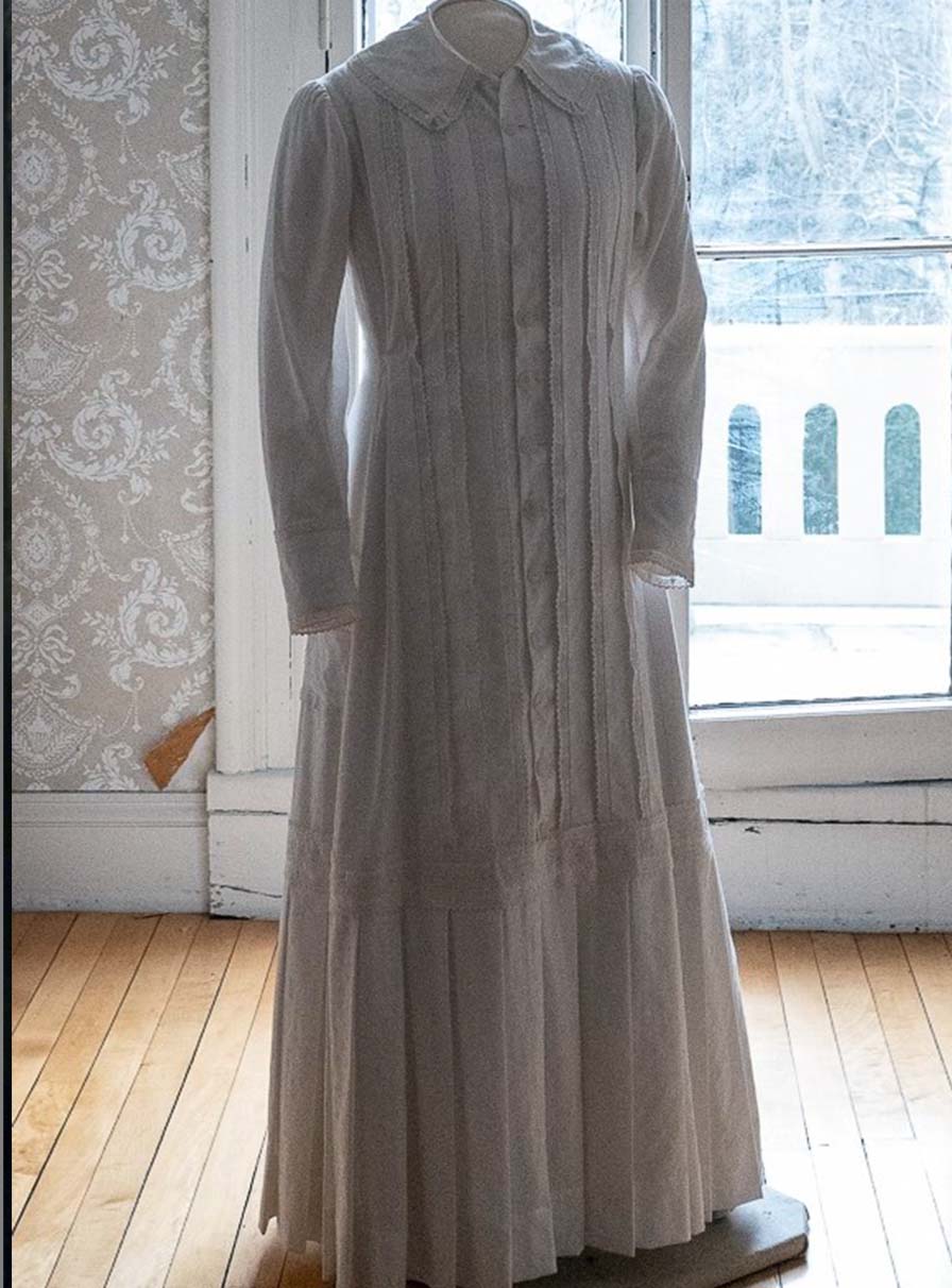 Front of a white cotton dress on mannequin in front of a large window. The bottom is finished with stiff box pleats.